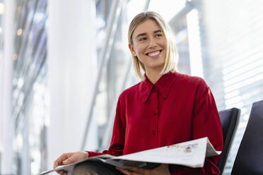 Lächelnde junge Geschäftsfrau mit Zeitung im Wartebereich sitzend - DIGF09000