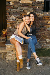 Zwei fröhliche Frauen mit einer Mappe umarmen sich an einem Steinhaus - MPPF00353