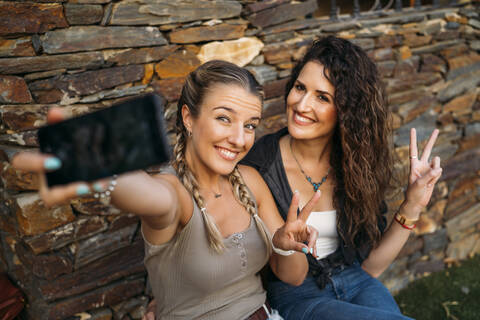 Zwei glückliche Frauen sitzen an einem Steinhaus und machen ein Selfie, lizenzfreies Stockfoto