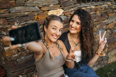 Zwei glückliche Frauen sitzen an einem Steinhaus und machen ein Selfie - MPPF00355