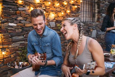 Happy couple playing ukulele outdoors at a stone house at dusk - MPPF00364