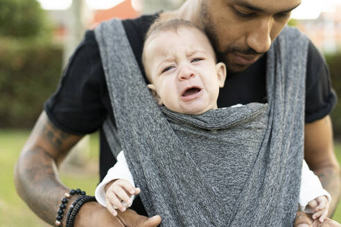 Junger Vater trägt sein weinendes Baby in einer Babytrage - ERRF02210