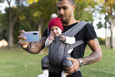 Young father carrying baby son in a baby sling, taking smartphone selfie - ERRF02213