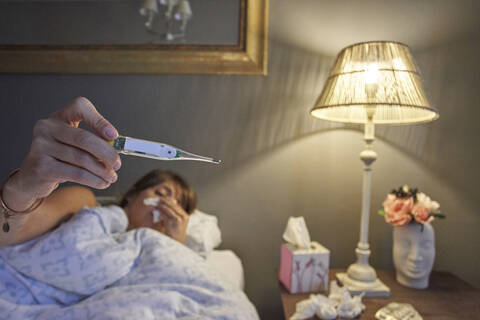 Kranke Frau, die im Bett liegt und ihre Temperatur mit dem Thermometer überprüft, lizenzfreies Stockfoto