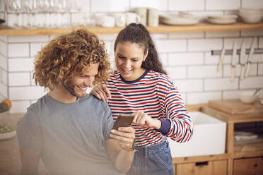 Young woman showing smartphone to man in the kitchen at home - MCF00384