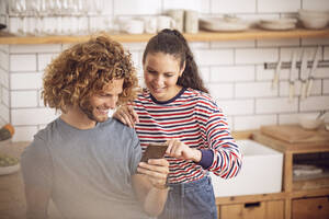 Junge Frau zeigt dem Mann in der Küche zu Hause ihr Smartphone - MCF00384