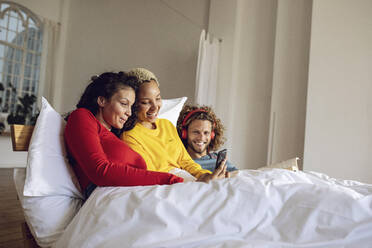 Happy friends relaxing in bed at home with smartphone - MCF00386