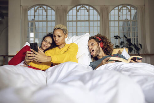 Glückliche Freunde haben Spaß im Bett zu Hause mit Smartphone - MCF00388