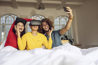Happy friends having fun with VR glasses and smartphone in bed at home - MCF00396