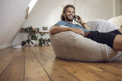 Lächelnder junger Mann entspannt sich in einem Sitzsack zu Hause und telefoniert, lizenzfreies Stockfoto