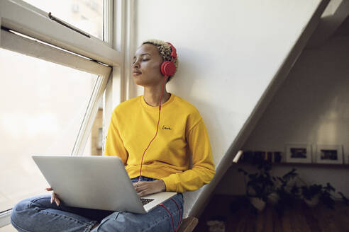 Junge Frau sitzt zu Hause am Fenster mit Laptop und Kopfhörern - MCF00407