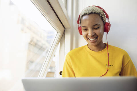 Glückliche junge Frau, die mit Laptop und Kopfhörern zu Hause am Fenster sitzt, lizenzfreies Stockfoto