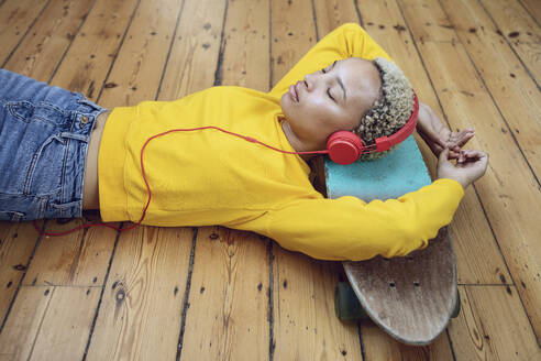 Young woman lying on skateboard on the floor at home listening to music - MCF00411