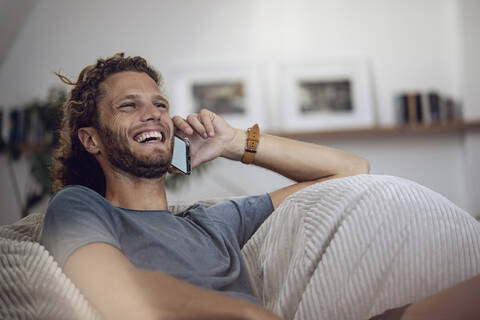 Lachender junger Mann, der sich zu Hause im Sitzsack entspannt und telefoniert, lizenzfreies Stockfoto