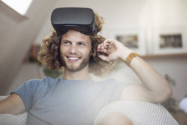 Portrait of smiling young man wearing VR glasses at home - MCF00419