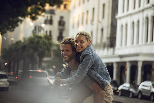 Happy young man carrying girlfriend piggyback in the city - MCF00426