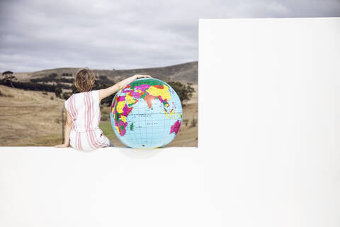 Kleines Mädchen an der Wand sitzend, den Arm um einen aufblasbaren Globus gelegt, Rückansicht, lizenzfreies Stockfoto