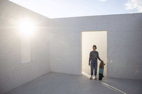 Girl with skateboard standing in empty, sunlit space - MCF00528