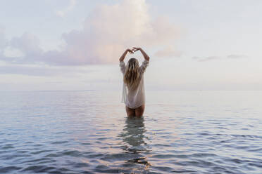 Rückansicht einer jungen Frau mit weißer Bluse im Meer bei Sonnenuntergang - AFVF04351