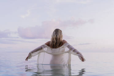 Rückansicht einer jungen Frau mit weißer Bluse im Meer bei Sonnenuntergang - AFVF04353