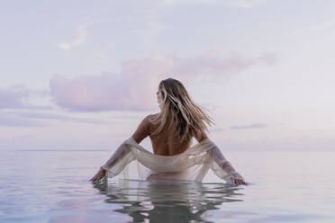 Rear view of young woman wearing white blouse inside sea during sunset - AFVF04354