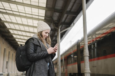 Lächelnde junge Frau mit Rucksack steht auf dem Bahnsteig und schaut auf ihr Handy, Vilnius, Litauen - AHSF01590