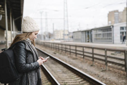 Lächelnde junge Frau mit Rucksack, die auf einem Bahnsteig steht und ein Mobiltelefon benutzt - AHSF01591