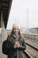 Portrait of smiling young woman with backpack and smartphone standing on platform - AHSF01592