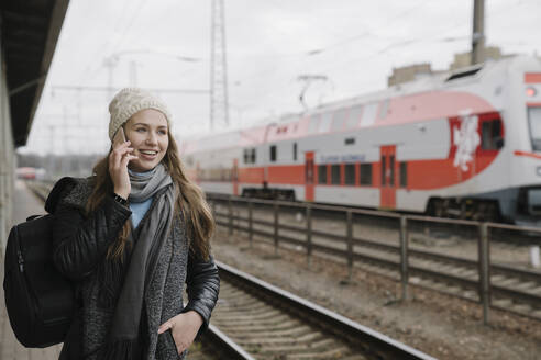 Porträt einer lächelnden jungen Frau, die am Telefon auf dem Bahnsteig wartet, Vilnius, Litauen - AHSF01594