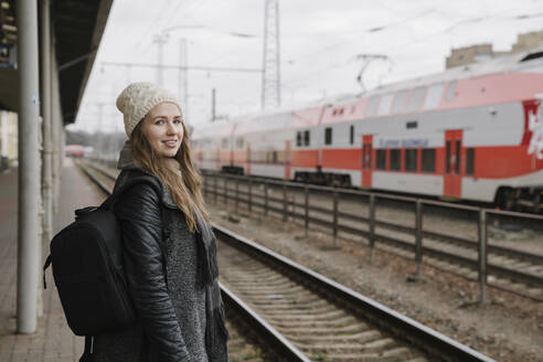 Porträt einer lächelnden jungen Frau mit Rucksack, die auf dem Bahnsteig wartet, Vilnius, Litauen - AHSF01595