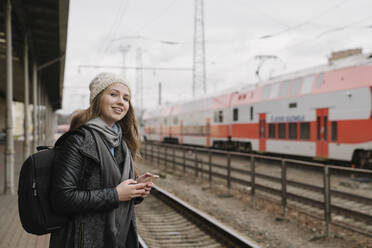 Porträt einer lächelnden jungen Frau mit Rucksack und Smartphone, die auf dem Bahnsteig wartet, Vilnius, Litauen - AHSF01596
