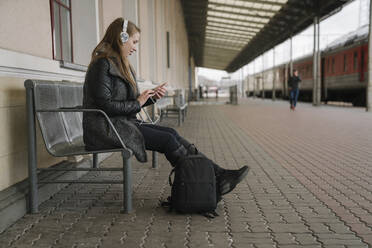 Lächelnde junge Frau mit Rucksack auf dem Bahnsteig sitzend, Smartphone und Kopfhörer benutzend, Vilnius, Litauen - AHSF01598