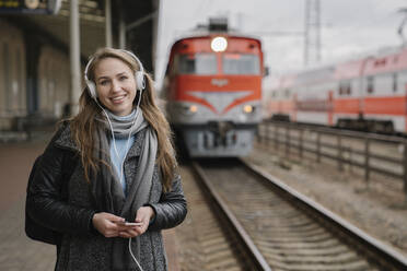 Portrait of happy woman standing on platform using smartphone and headphones, Vilnius, Lithuania - AHSF01599