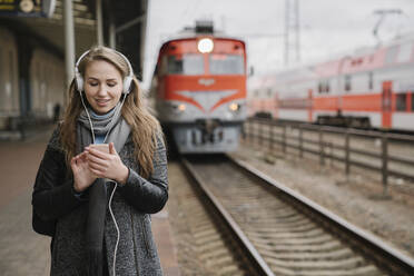 Smiling young woman standing on platform using smartphone and headphones, Vilnius, Lithuania - AHSF01600