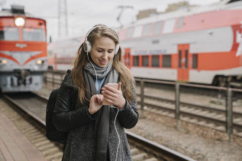 Lächelnde junge Frau auf dem Bahnsteig mit Smartphone und Kopfhörern, Vilnius, Litauen, lizenzfreies Stockfoto