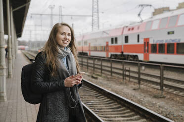 Smiling woman waiting on platform using smartphone and earphones, Vilnius, Lithuania - AHSF01602