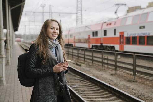 Lächelnde Frau, die mit Smartphone und Kopfhörern auf dem Bahnsteig wartet, Vilnius, Litauen - AHSF01602