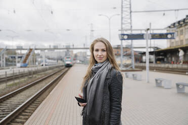 Porträt einer lächelnden jungen Frau mit Smartphone auf einem Bahnsteig - AHSF01603