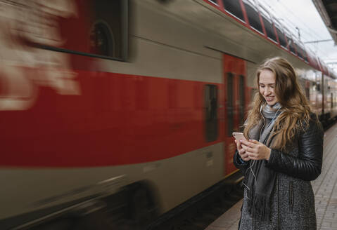 Smiling young woman standing on platform using smartphone and earphones, Vilnius, Lithuania - AHSF01604