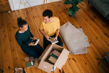 Smiling couple removing novels from box in living room at new home - MASF14748