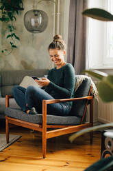 Mittlere erwachsene Frau lächelt, während sie auf einem Sessel zu Hause ein Mobiltelefon benutzt - MASF14760