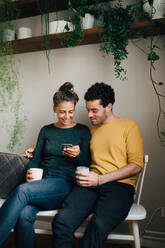 Lächelnder Freund und Freundin schauen auf ihr Handy, während sie im Wohnzimmer Kaffee trinken - MASF14781