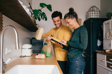 Smiling man removing food from bag while girlfriend showing digital tablet at kitchen counter - MASF14792