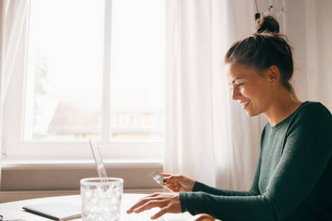 Seitenansicht einer lächelnden Frau mit Kreditkarte und Laptop im Wohnzimmer, lizenzfreies Stockfoto