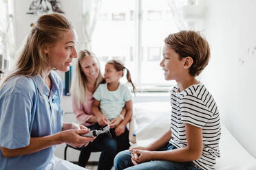 Lächelnde Kinderärztin mit Otoskop im Gespräch mit einem Jungen, während die Familie im Hintergrund in einer medizinischen Klinik sitzt - MASF14892
