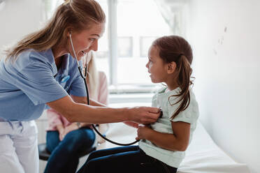 Kinderärztin, die den Herzschlag eines Mädchens mit dem Stethoskop untersucht, während die Mutter im Hintergrund in der Klinik sitzt - MASF14915