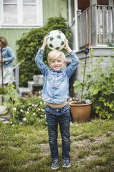 Lächelnder Junge in voller Länge, der einen Fußball auf dem Kopf hält, während er im Garten steht - MASF14925