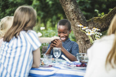Lächelnder Junge schaut auf sein Handy, während er mit Freunden am Esstisch im Hinterhof sitzt - MASF14935