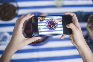 Ausgeschnittene Hand eines Mädchens mit Smartphone, das einen Jungen beim Dessert am Tisch fotografiert - MASF14946