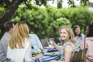 Lächelnde Frau hält ein Weinglas, während sie mit Freunden auf einer Gartenparty sitzt - MASF14956